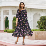 Woman standing in a black floral print A-line midi dress with sleeves, smiling in front of white palace backdrop.