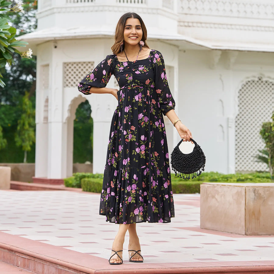 Model wearing black dress with sleeves with floral print and holding black bag.
