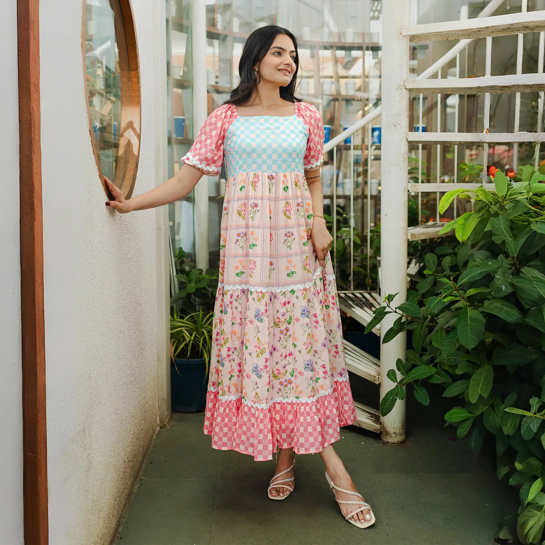 Model posing sideways near a window in a floral A-line maxi dress, styled for vacation outings.